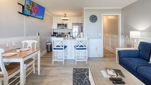 Een moderne keuken met witte kasten, een magnetron en een koffieapparaat. Er is een eethoek met een houten tafel en witte stoelen. In de woonkamer staat een blauwe bank en een klein vloerkleed.