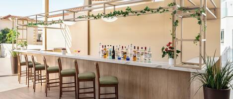 A bar area with wooden chairs, a counter with bottles, and a pergola with greenery.