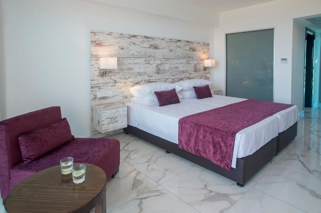 Quarto de hotel moderno com uma cama grande, um sofá vermelho, uma mesa pequena com copos e uma cabeceira de madeira.