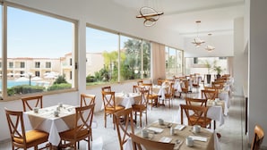 Zona pranzo con tavoli apparecchiati, ampie finestre e vista su piscina e edifici.