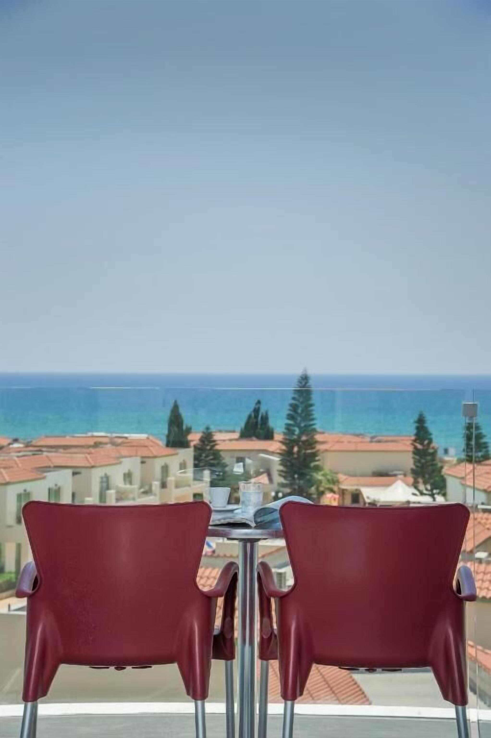 Un balcón con dos sillas rojas y una mesa, con vista a la costa, edificios y árboles.