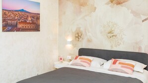 A hotel room with a bed, pillows, a nightstand, a wall-mounted lamp, and a framed cityscape picture.