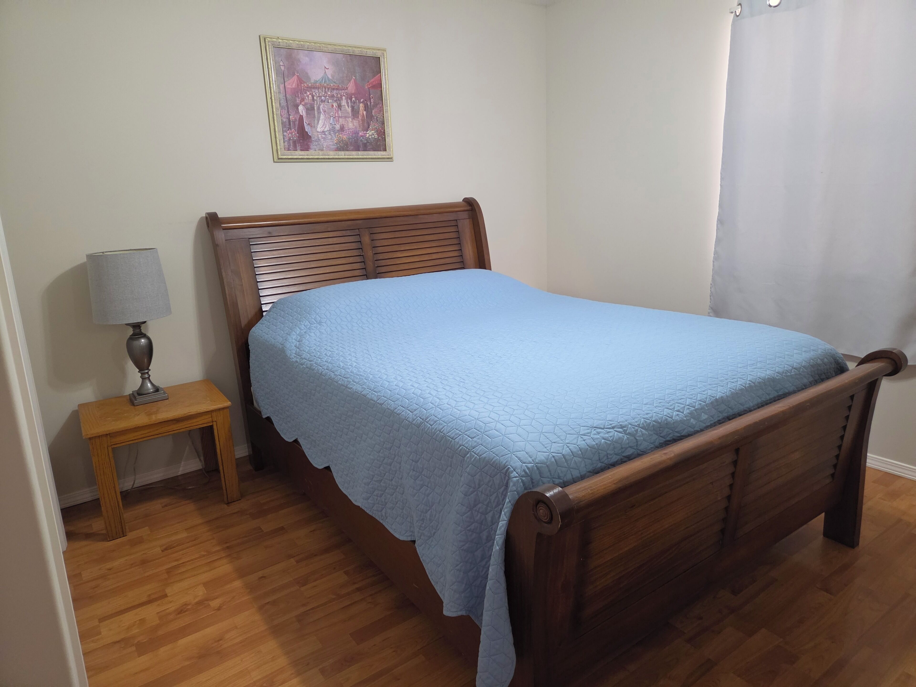 Quarto com cama de madeira, mesa de cabeceira com abajur, quadro na parede e piso de madeira.