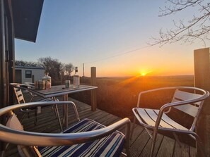 A deck with chairs and a table, overlooking a sunset. - Elizabeth Carne (No Man's Land)
