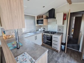 A compact kitchen with a sink, stove, oven, and a window with blinds. - Elizabeth Carne (No Man's Land)