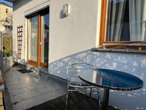 Un balcon avec une table et une chaise, une porte-fenêtre coulissante, une échelle en bois et une fenêtre avec des rideaux.