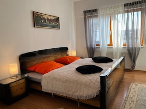 Ein Schlafzimmer mit Bett, Nachttischen, einem Gemälde und Blick durch leichte Vorhänge.