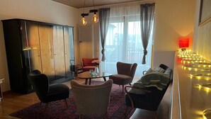 Een woonkamer met een zwarte kast, een rode stoel, een beige fauteuil, een kamerkleed met patroon en een tafel met een bloevas.