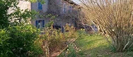 A rustic building with a stone facade, a small garden with grass and a tree, and a fence in the background.