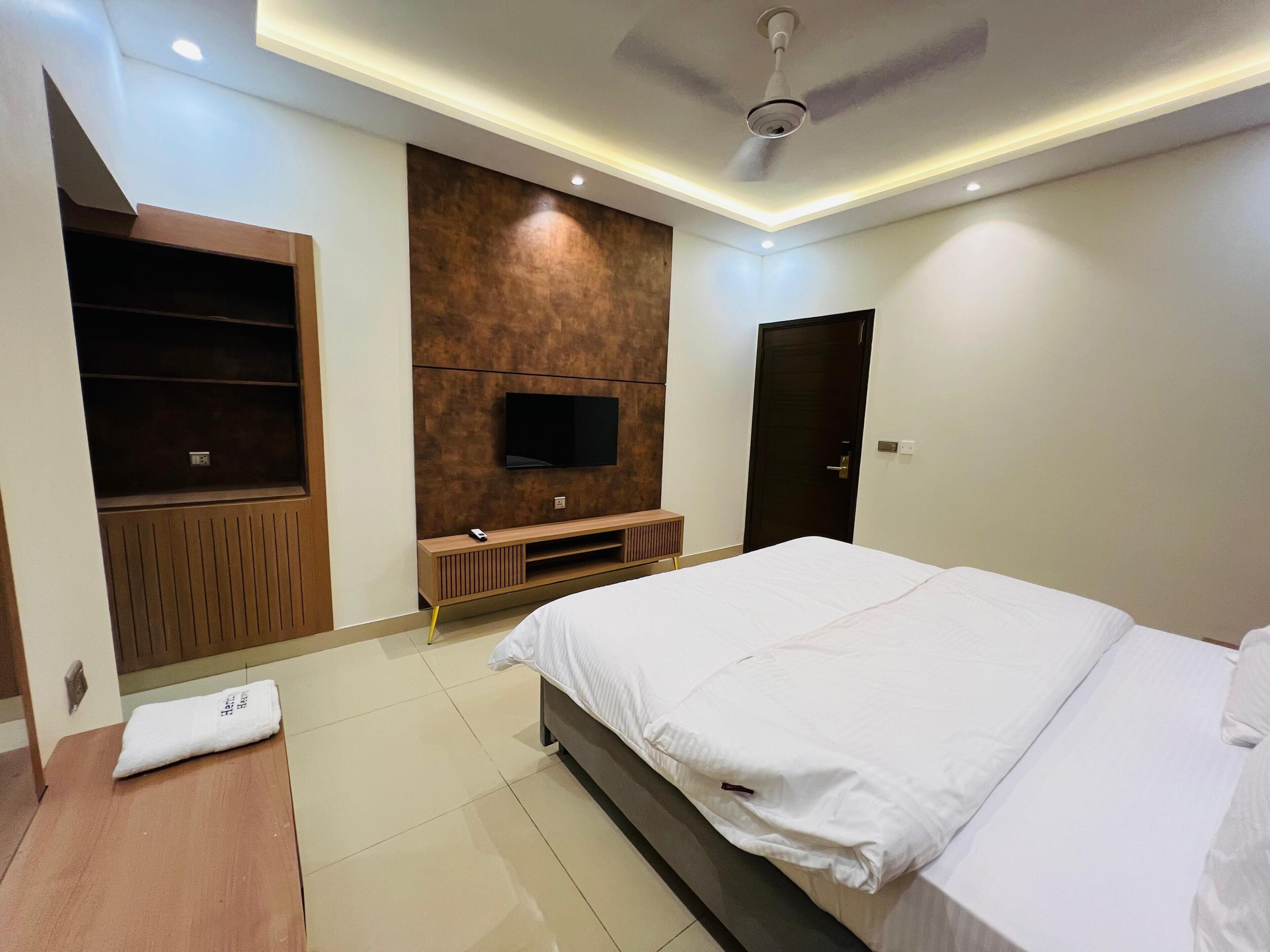 A modern hotel room with a large bed, a TV mounted on the wall, a wooden cabinet, and a ceiling fan.