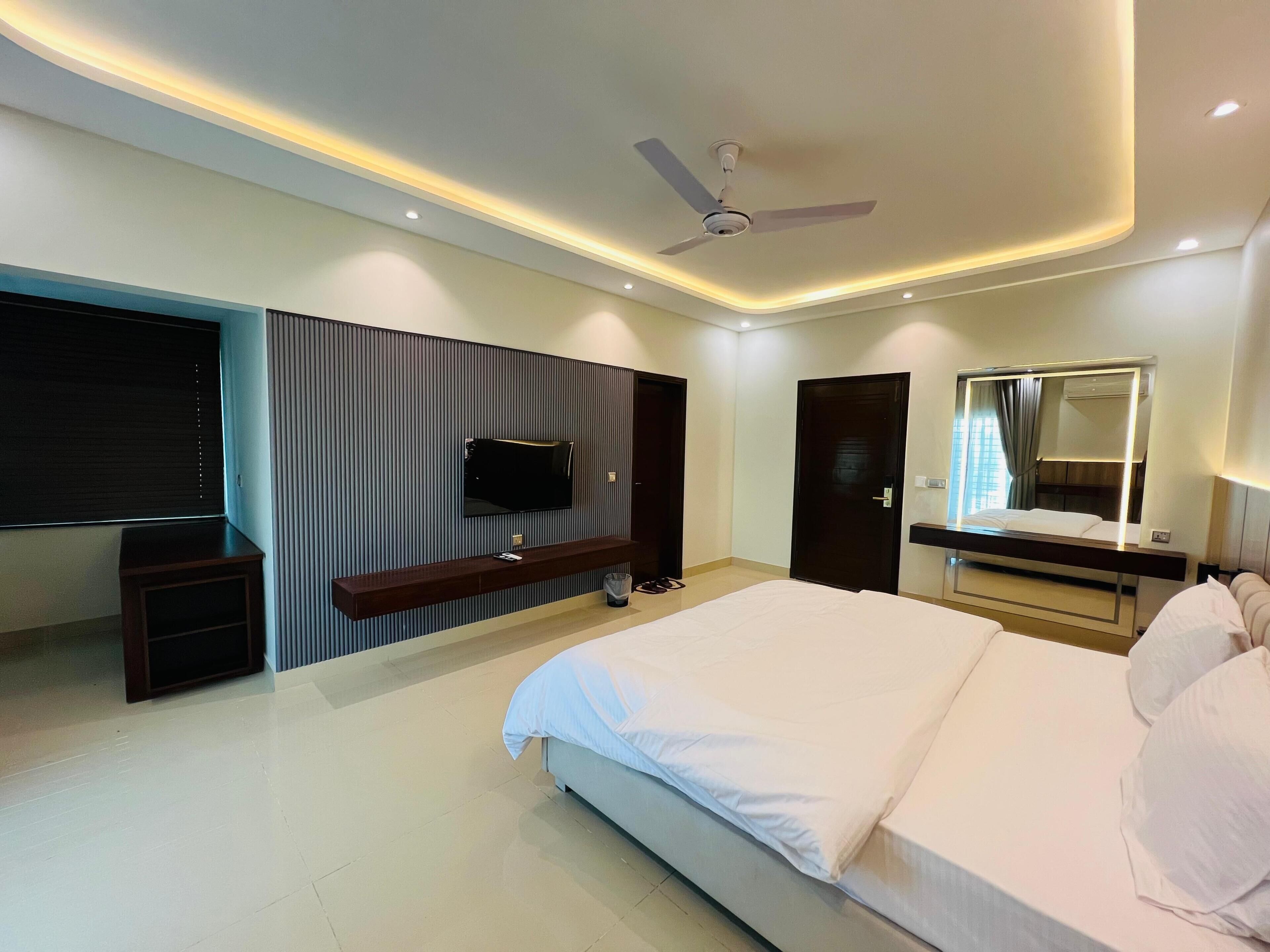 A modern hotel room with a large bed, a flat-screen TV mounted on the wall, a ceiling fan, and a large mirror above the bed.