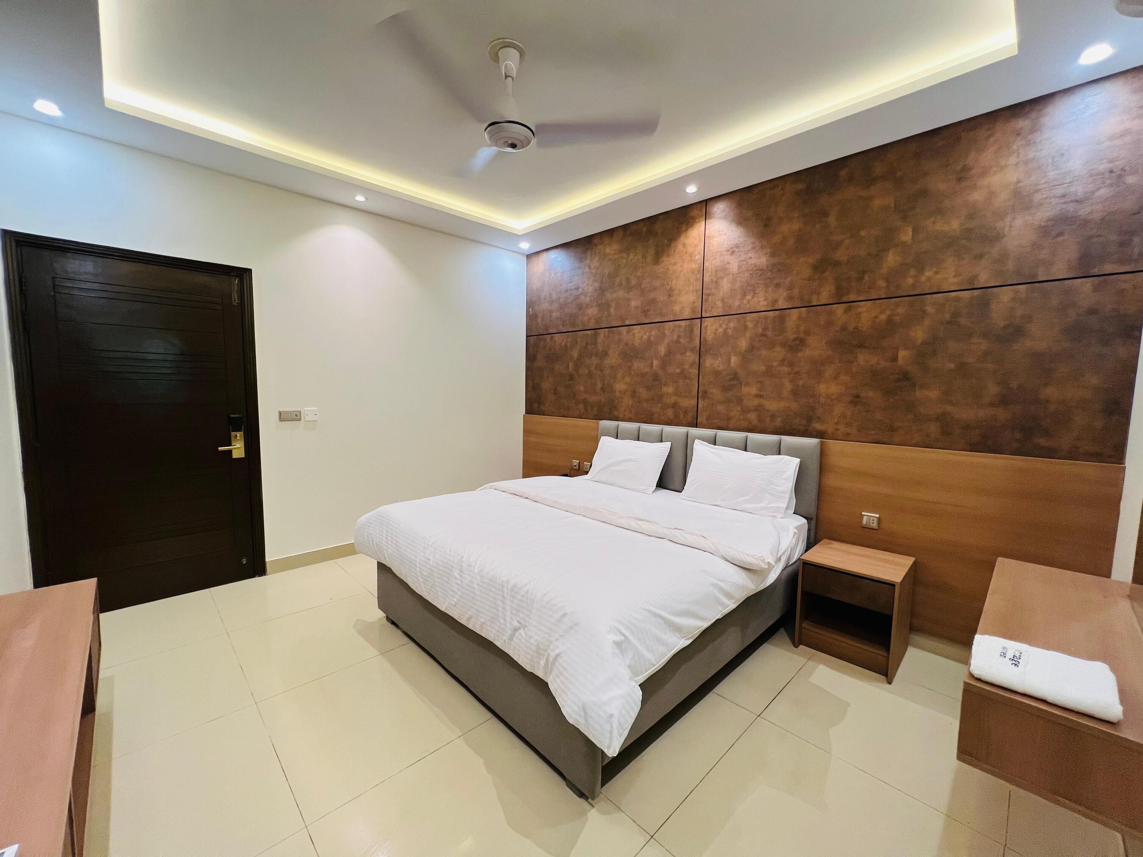 A modern hotel room with a large bed, bedside tables, a wooden headboard, and recessed ceiling lighting.