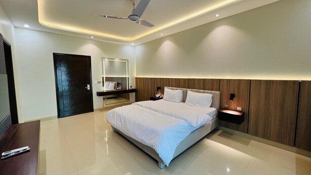 A modern bedroom with a large bed, a ceiling fan, recessed lighting, a wooden headboard, and a nightstand with a lamp. - Heritage Heaven (Karachi)