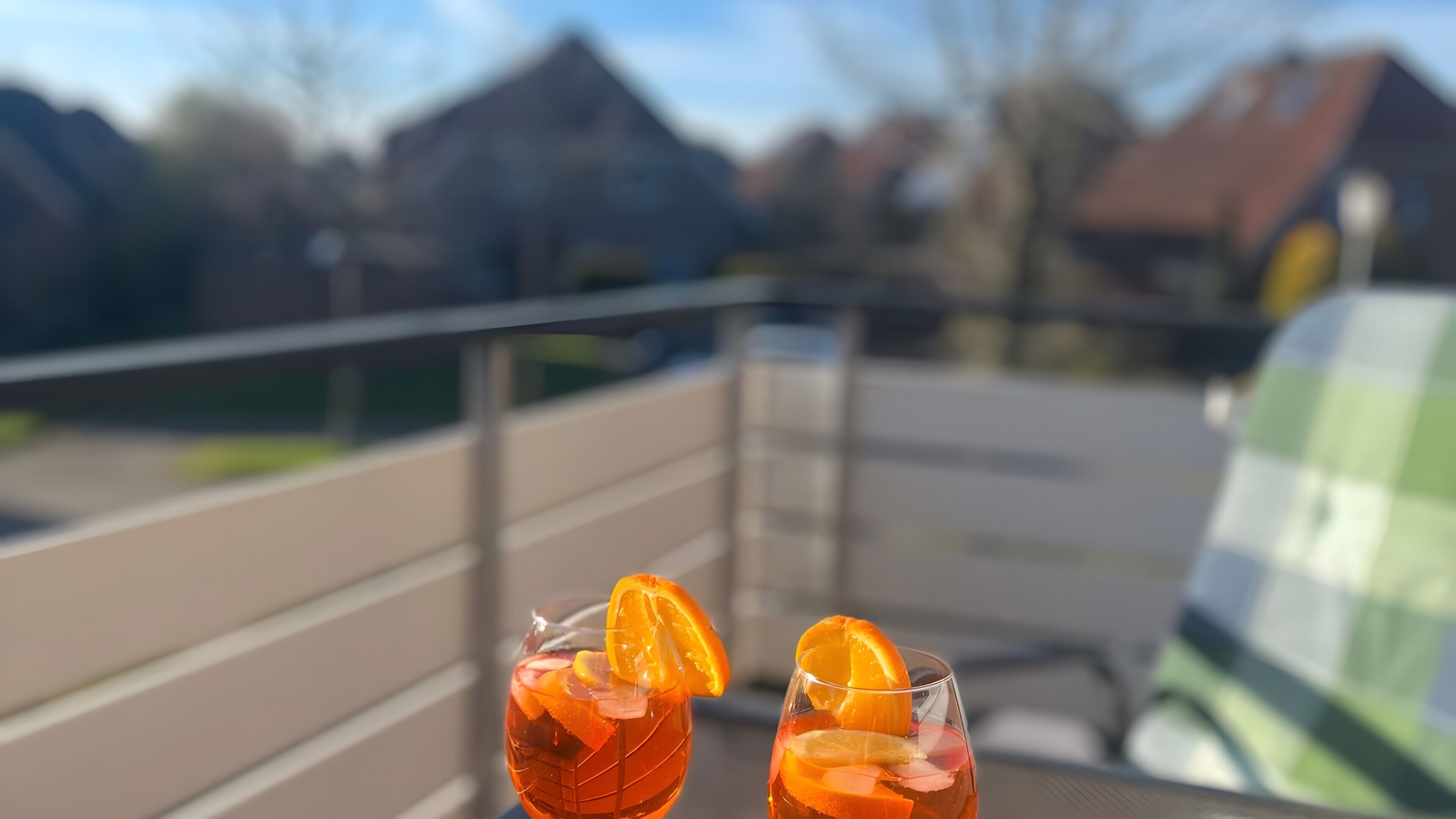 Two glasses of a beverage with orange slices on a balcony with a railing, overlooking a residential area.