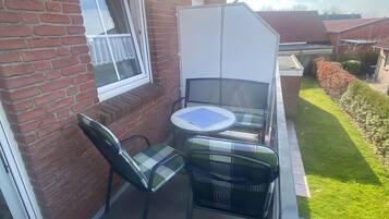 Une petite terrasse extérieure avec une table et des chaises, une fenêtre ornée d’un rideau blanc et un mur en briques.