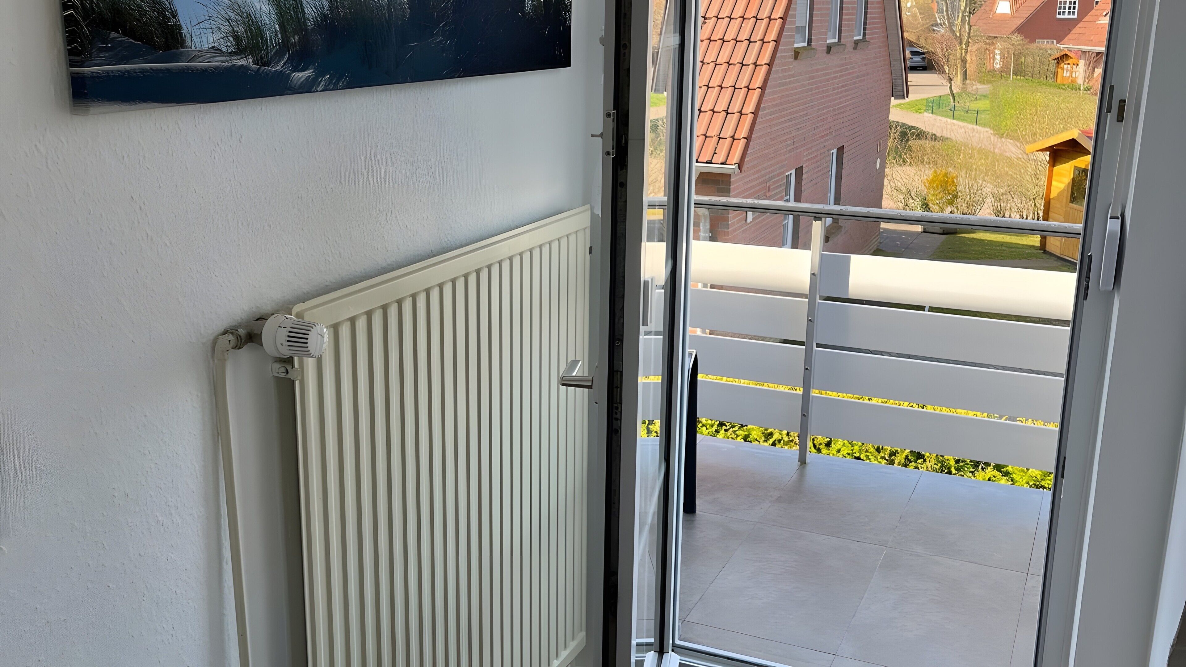 Un balcón con vista a una zona residencial, un radiador blanco y una puerta con un panel de vidrio.