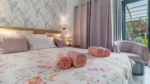 Ein Schlafzimmer mit floralem Bettbezug, einem Holzkopfteil, einem Nachttisch mit Lampe, einem Sessel und Blick auf Grün durch ein Fenster.