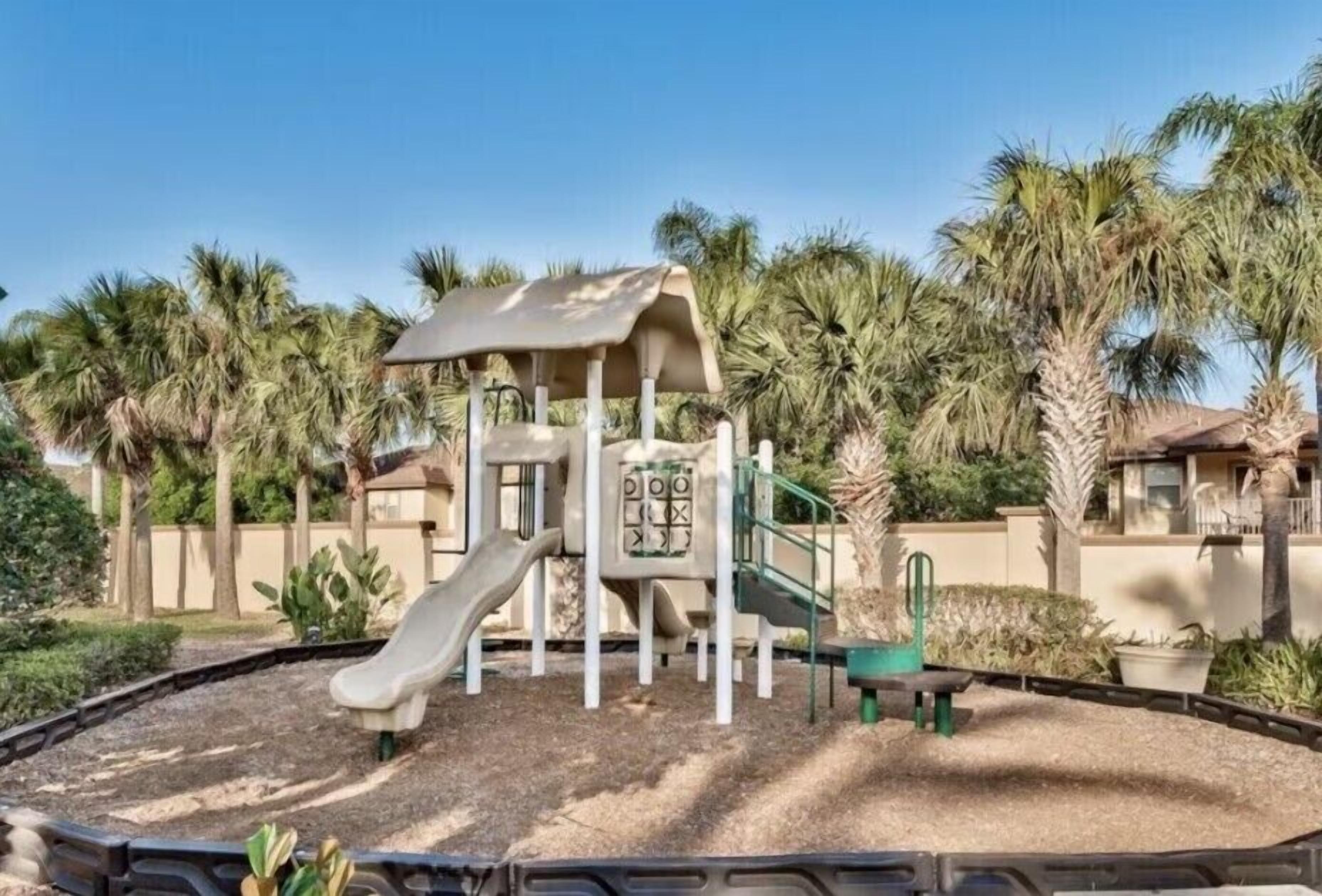 A playground with a slide and climbing structures, surrounded by palm trees and a fence.