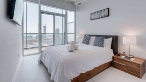 A modern bedroom with a large bed, a nightstand, a TV mounted on the wall, and a city view through large windows.