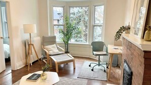 A room with a fireplace, a large window, a standing lamp, a chair, a desk, and a plant.