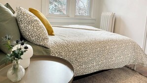 A bedroom with a bed, pillows, a bedside table, a vase, and a window.