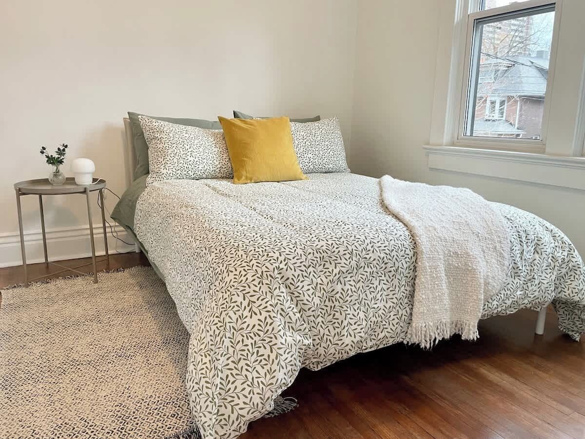 A bedroom with a patterned bedspread, a small bedside table with a vase, and a view through a window.