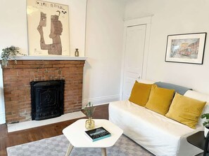 A living room with a brick fireplace, a white sofa with yellow cushions, a small white table with a book, and a framed picture on the wall. - Charming + Airy Roncy Apartment (Toronto)