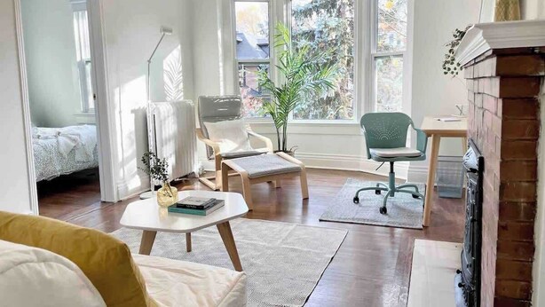 A room with a bed, two chairs, a small table, a fireplace, and large windows.
