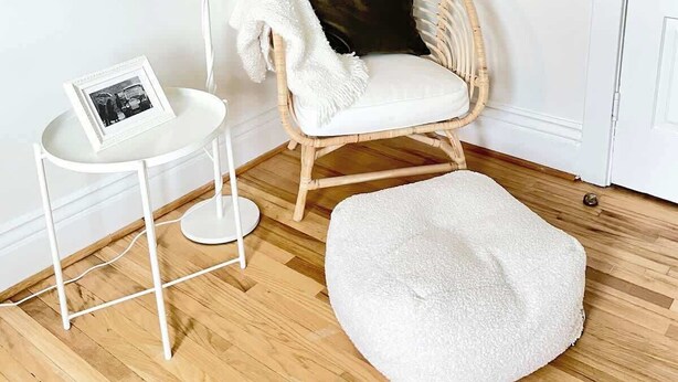 Una habitación de esquina con piso blanco, una lámpara blanca, una silla de mimbre con cojín, una mesita pequeña con un cuadro y un otoman blanco.