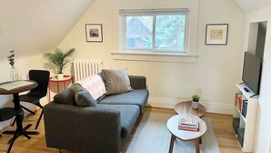 Un salón acogedor con un sofá gris, una mesita redonda, un mueble para televisión y una ventana con vistas a una casa.