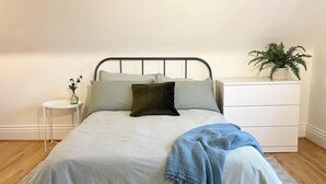Una cama con estructura metálica, una mesita de noche blanca con una planta y un aparador blanco con cajones.