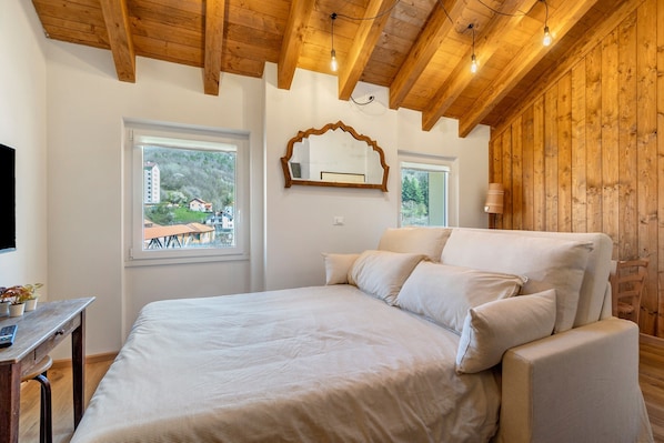 Une chambre à coucher comprenant un lit, un bureau, un miroir et une fenêtre donnant sur des immeubles et des arbres.