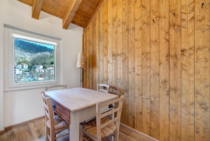 Une pièce aux murs lambrissés, avec une table et des chaises, une fenêtre donnant sur des maisons et des collines, et une lampe fixée au mur.
