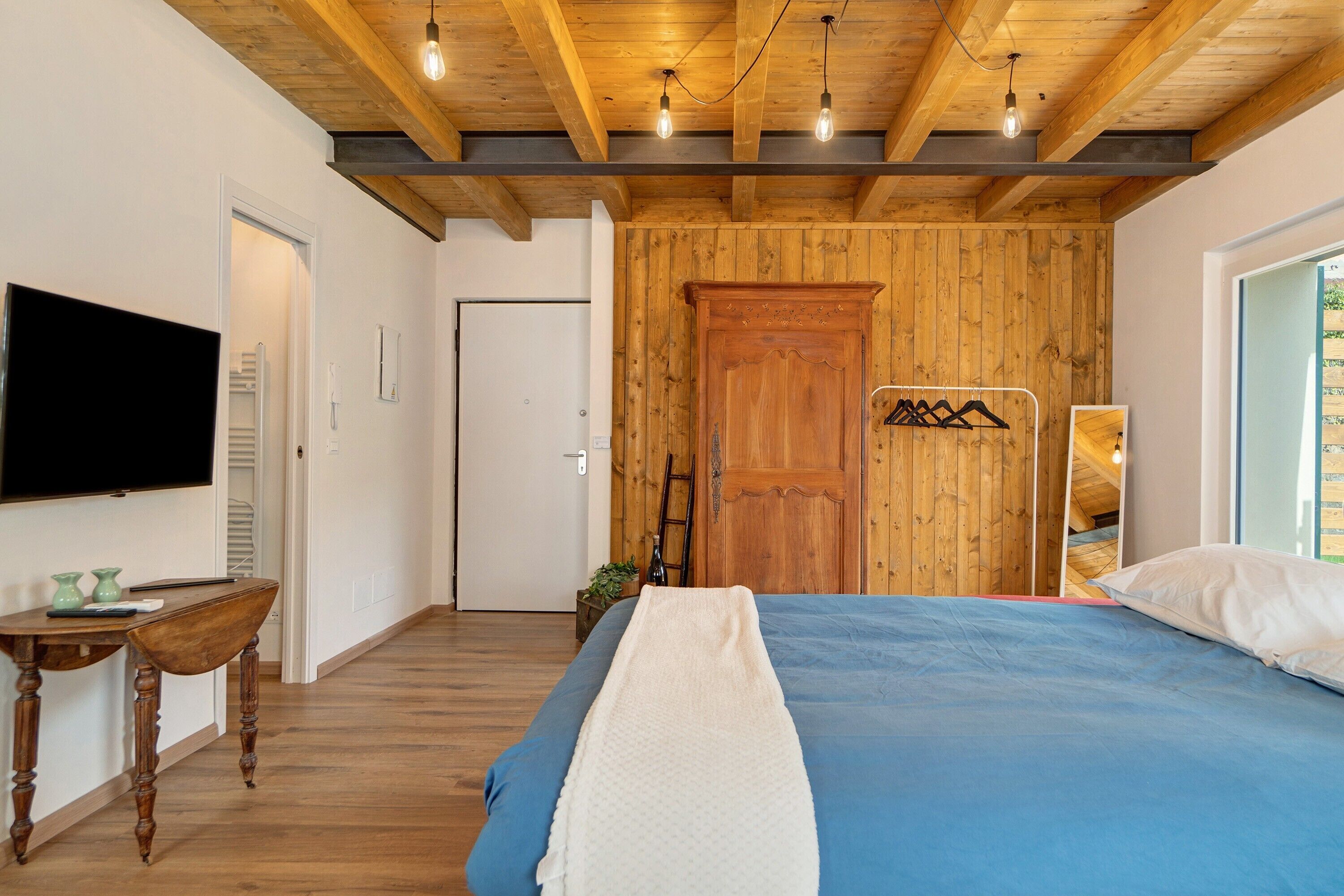 Ein Schlafzimmer mit Holzbalkendecke, einem Bett mit blauer Bettdecke, einer weißen Decke, einem Holzschrank und einem Fernseher an der Wand.