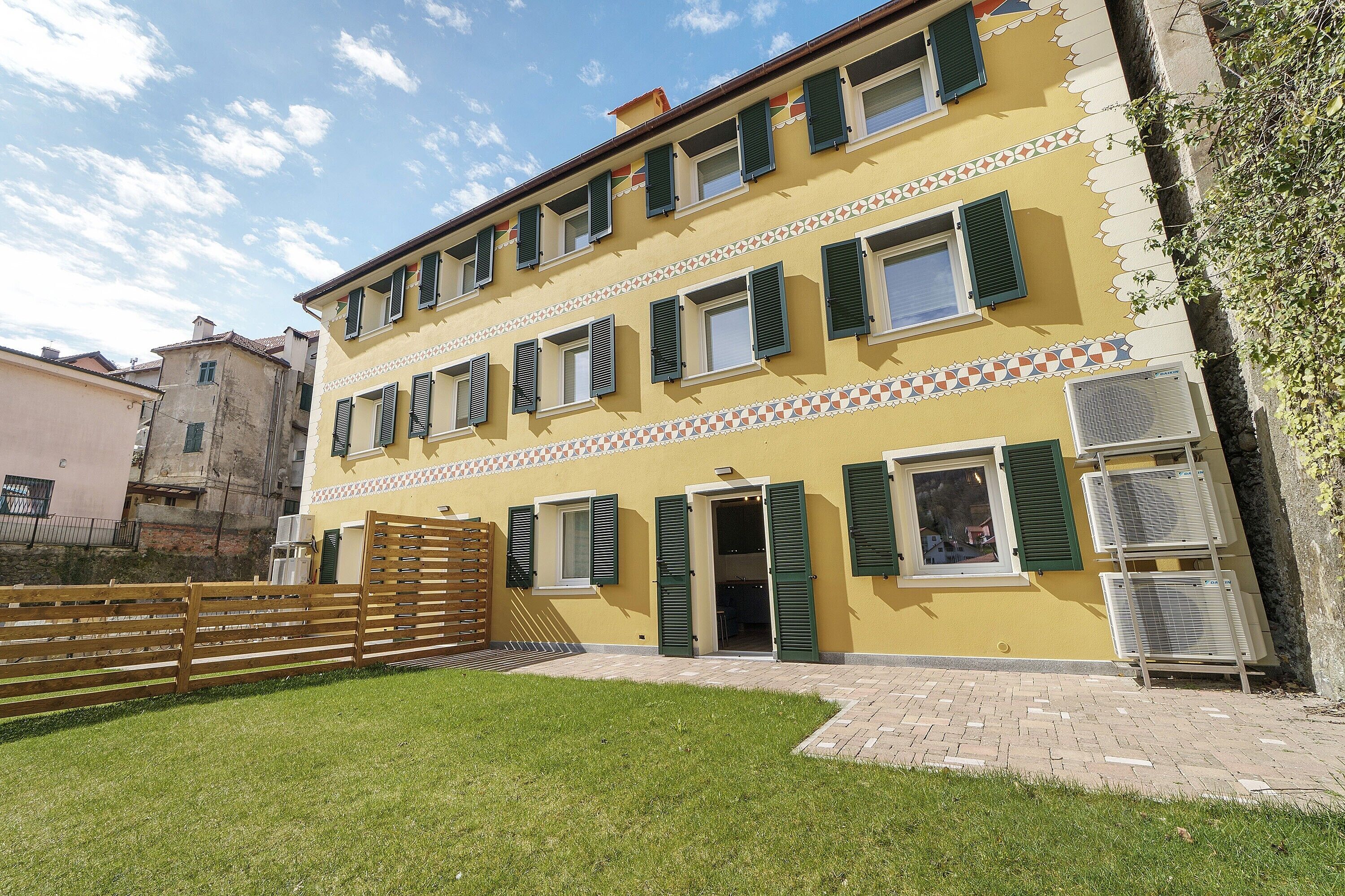 Un immeuble à plusieurs étages, doté d’une façade jaune, de volets verts et d’une cour pavée.