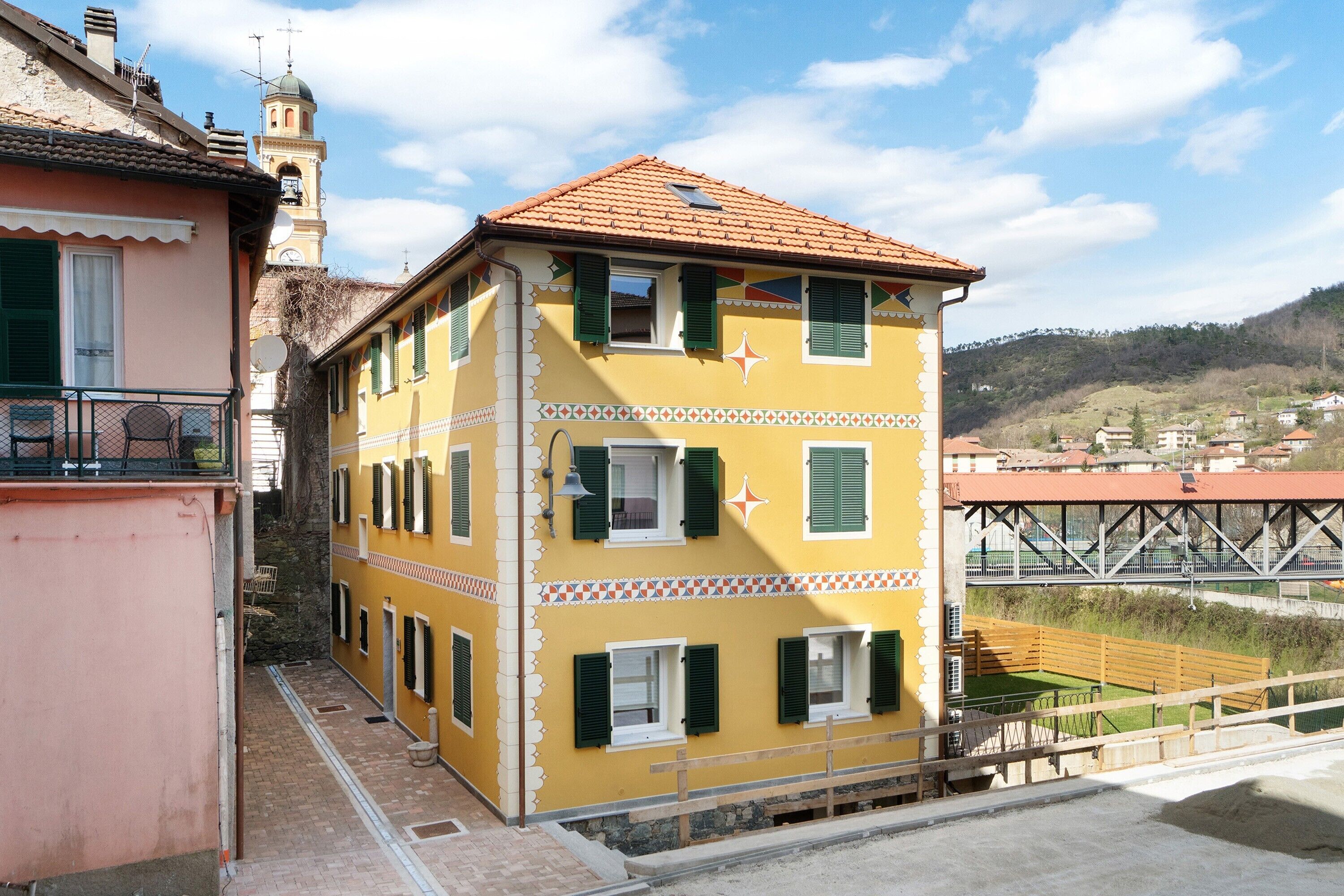 A yellow building with multiple windows and a balcony, adjacent to a pink building, with a bridge and hills in the background.