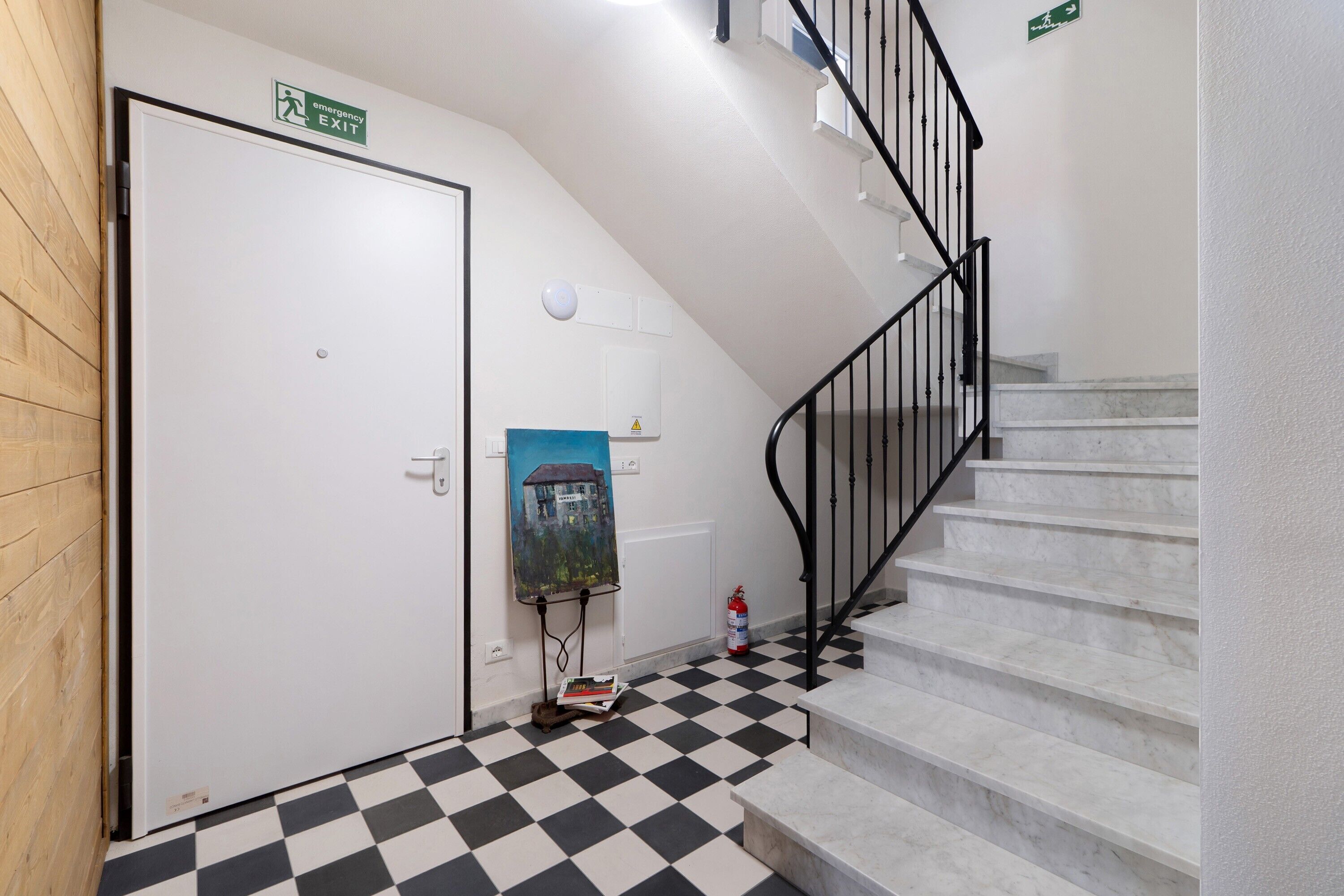 Eine Treppe mit schwarzer Geländer, eine weiße Tür mit einem Notausgangsschild, ein Schachbrettfußboden und ein Gemälde an der Wand.