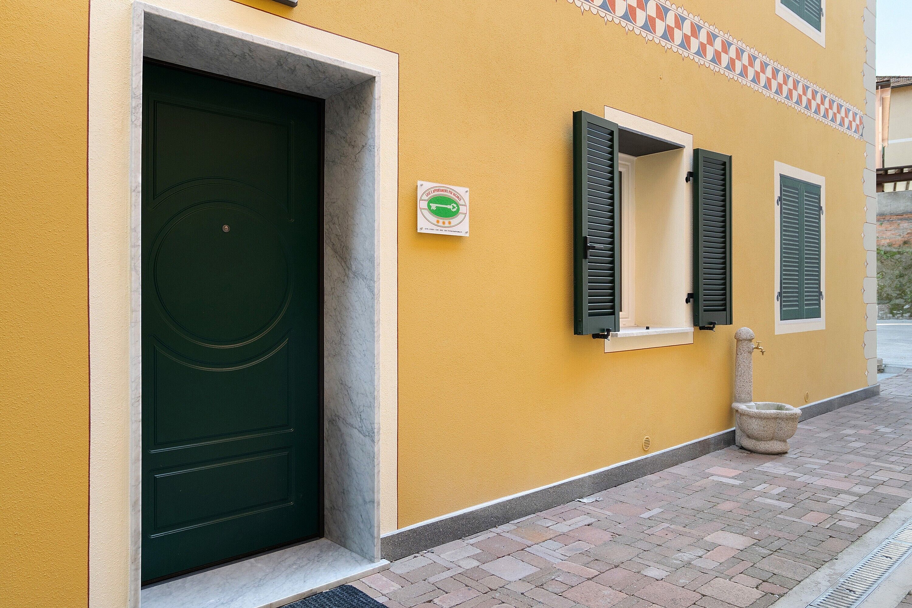 A building with a green door, a window with green shutters, and a stone water fountain on the sidewalk.