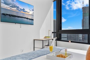 A modern room with a large window, a flat-screen TV, a small table with books, and a breakfast tray. - Miami Vacation Rentals - District 225 (Miami)