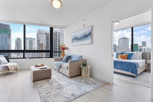 A modern living room with a sofa, ottoman, and a coffee table, a bed with a blue blanket, and a city view through large windows.