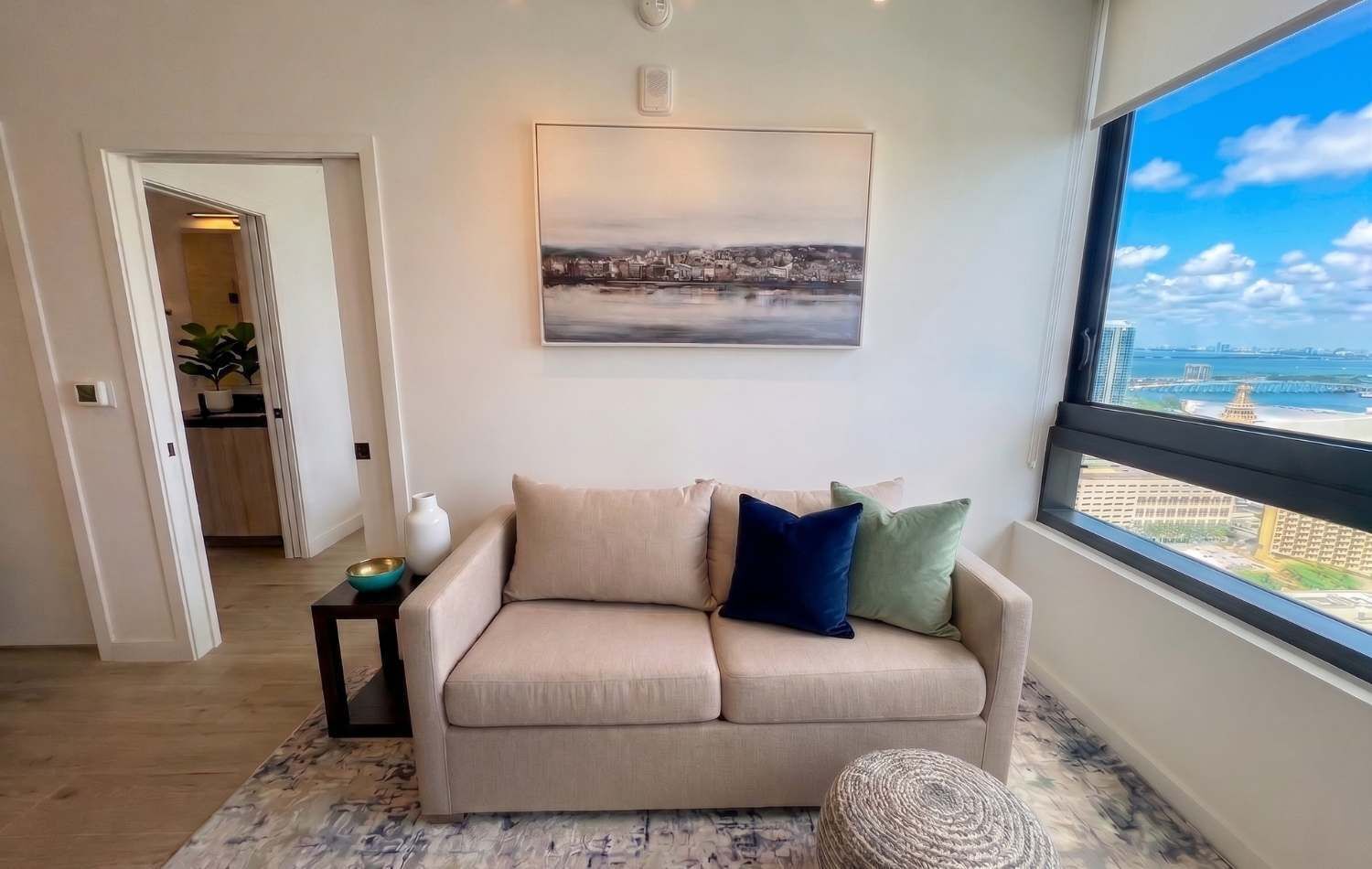 Un salón con un sofá color beige, una mesita auxiliar pequeña, un otoman tejido y un ventanal con vistas a la ciudad.