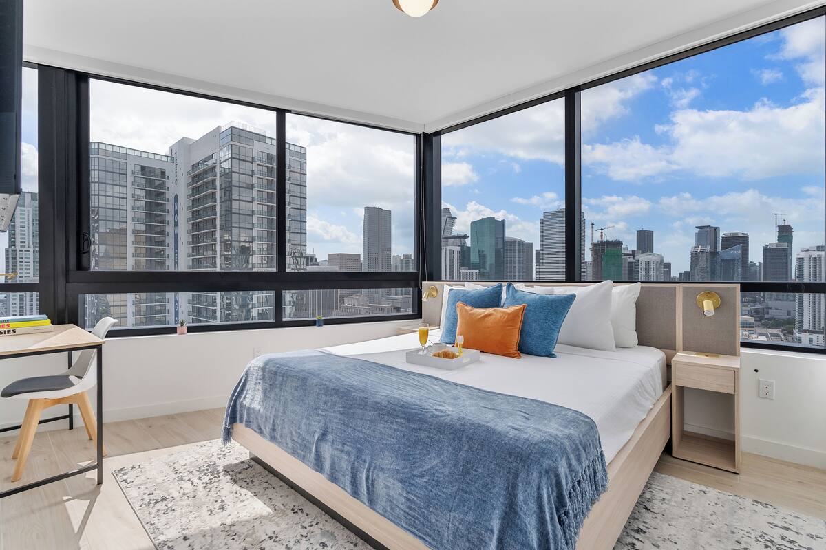Un dormitorio moderno con una cama grande, un escritorio y vistas a un horizonte urbano a través de amplios ventanales.