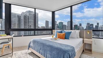 A modern bedroom with a large bed, a desk, and a view of a city skyline through large windows.