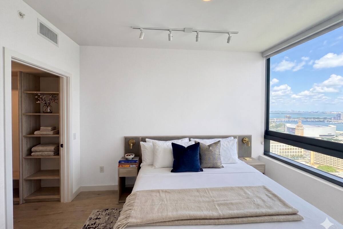Un dormitorio con cama, mesita de noche, armario, ventana con vista a la ciudad y lámpara de techo.
