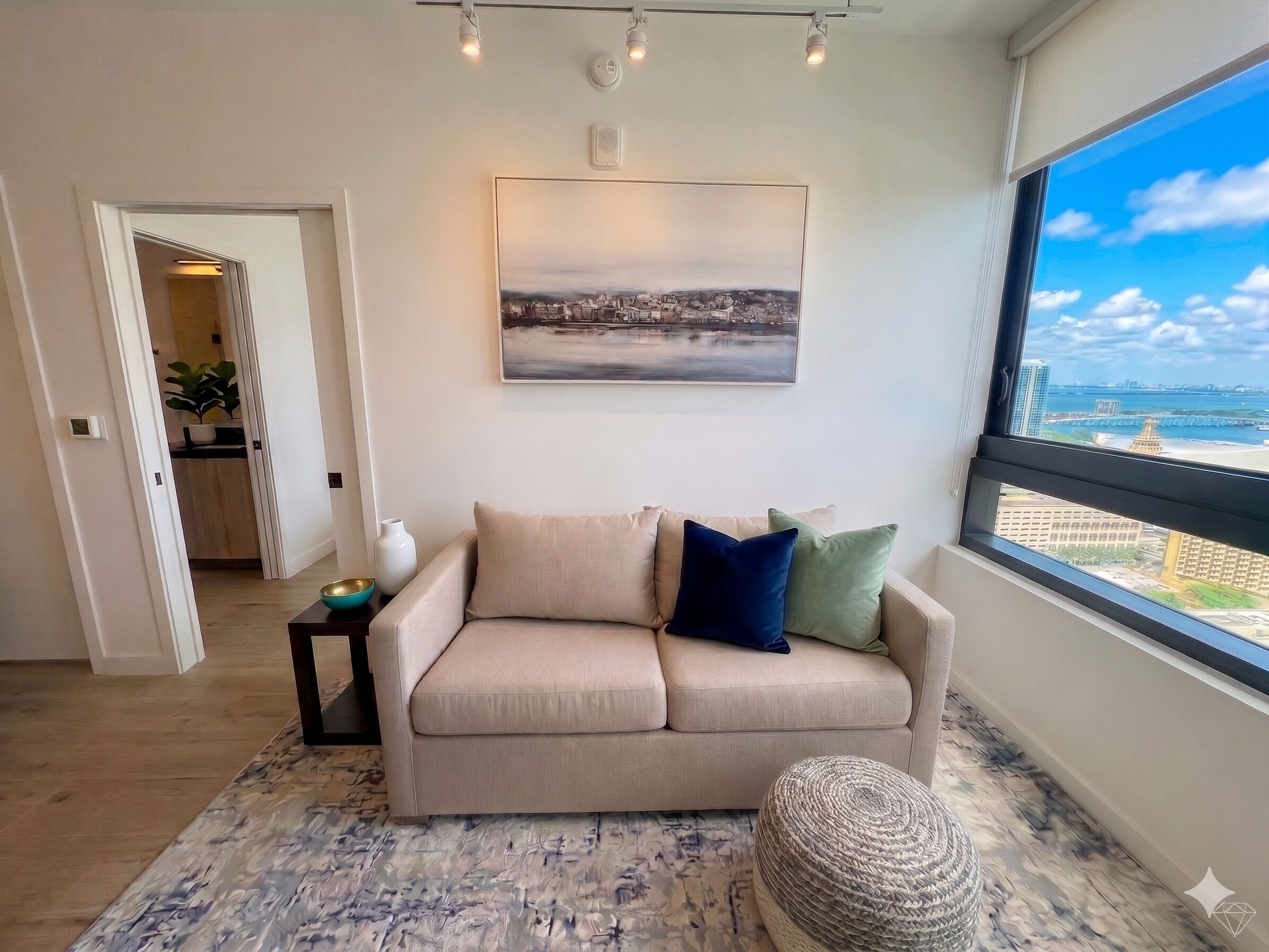 A living room with a sofa, a painting, a window with a view, and a small table with a bowl.