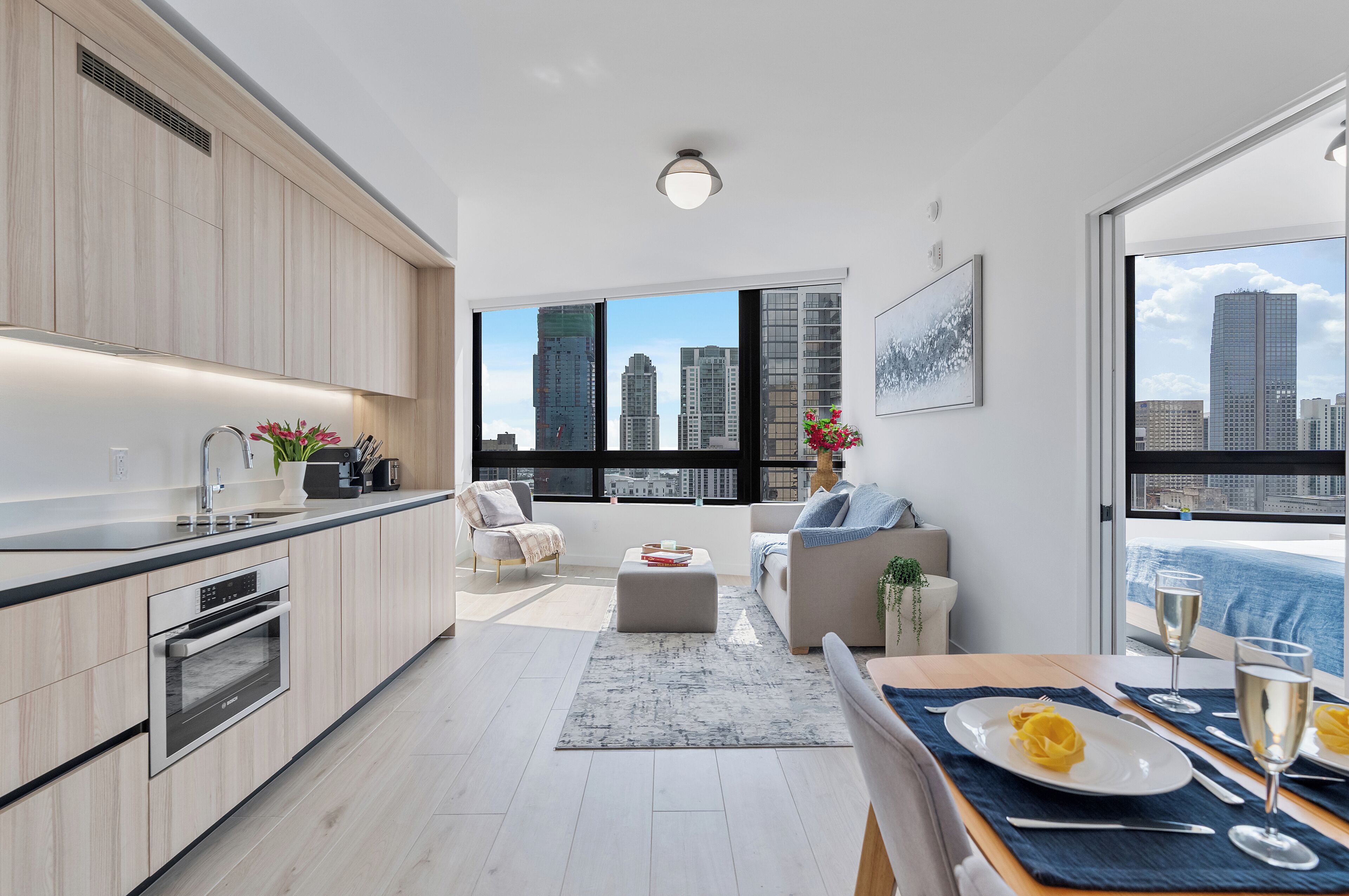 Une cuisine moderne offrant une vue sur les gratte-ciel de la ville, un coin repas avec une table dressée pour deux personnes, et un espace salon confortable avec un canapé et un fauteuil.
