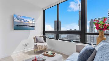 Uma sala de estar moderna com uma grande janela, vista para o horizonte da cidade, um espaço de estar aconchegante e uma pequena mesa com objetos decorativos.