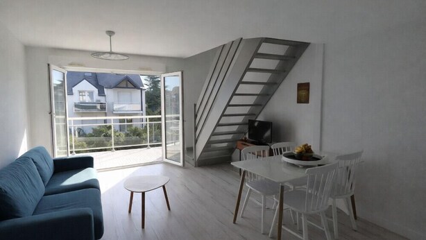 Uma sala de estar moderna com sofá, mesa de jantar com cadeiras, escada e uma janela grande com vista para casas.