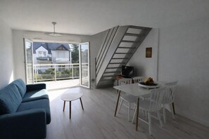 Een moderne woonkamer met een bank, een eettafel met stoelen, een trap en een groot raam met uitzicht op huizen.