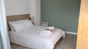 Uma cama bem arrumada com lençóis brancos e cabeceira de madeira, uma pequena mesa de cabeceira com abajur e um radiador na parede.